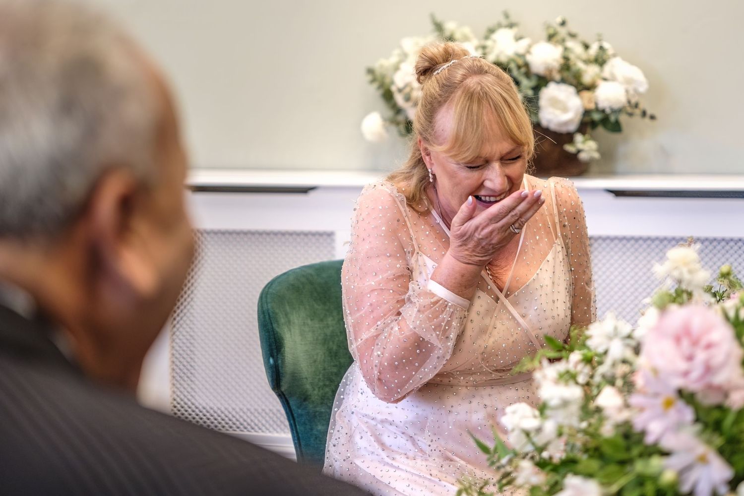 Emotional bride in lace dress covering mouth with hands, seated near pink floral arrangements at wedding ceremony.