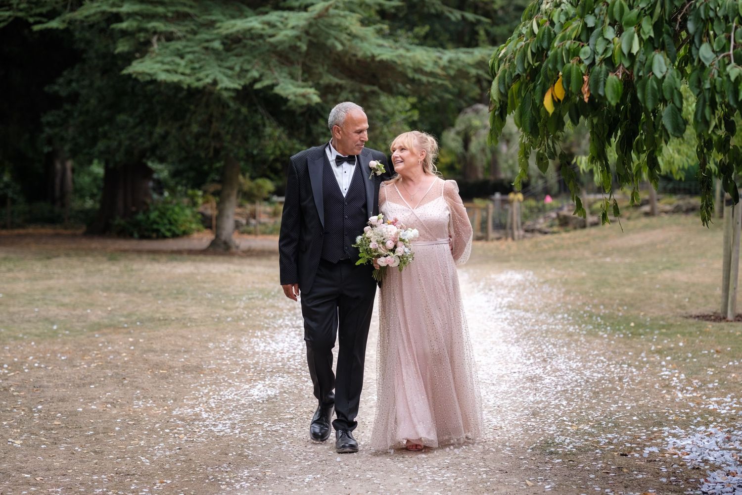 Elegant couple in wedding attire walking hand in hand along a tree-lined garden path, bride holding a bouquet.