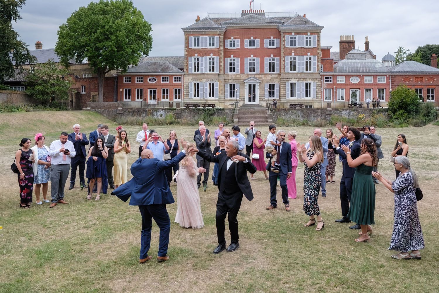 Wedding guests mingle on lawn before grand red brick Georgian manor house on sunny summer day.