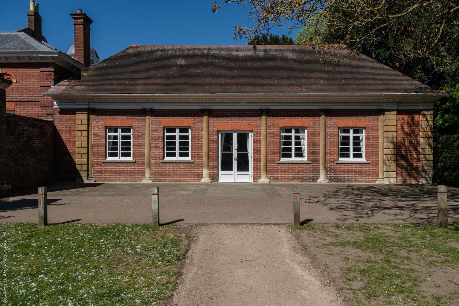 Historic red brick building with white-framed windows and a slate roof, surrounded by trees on a sunny day.
