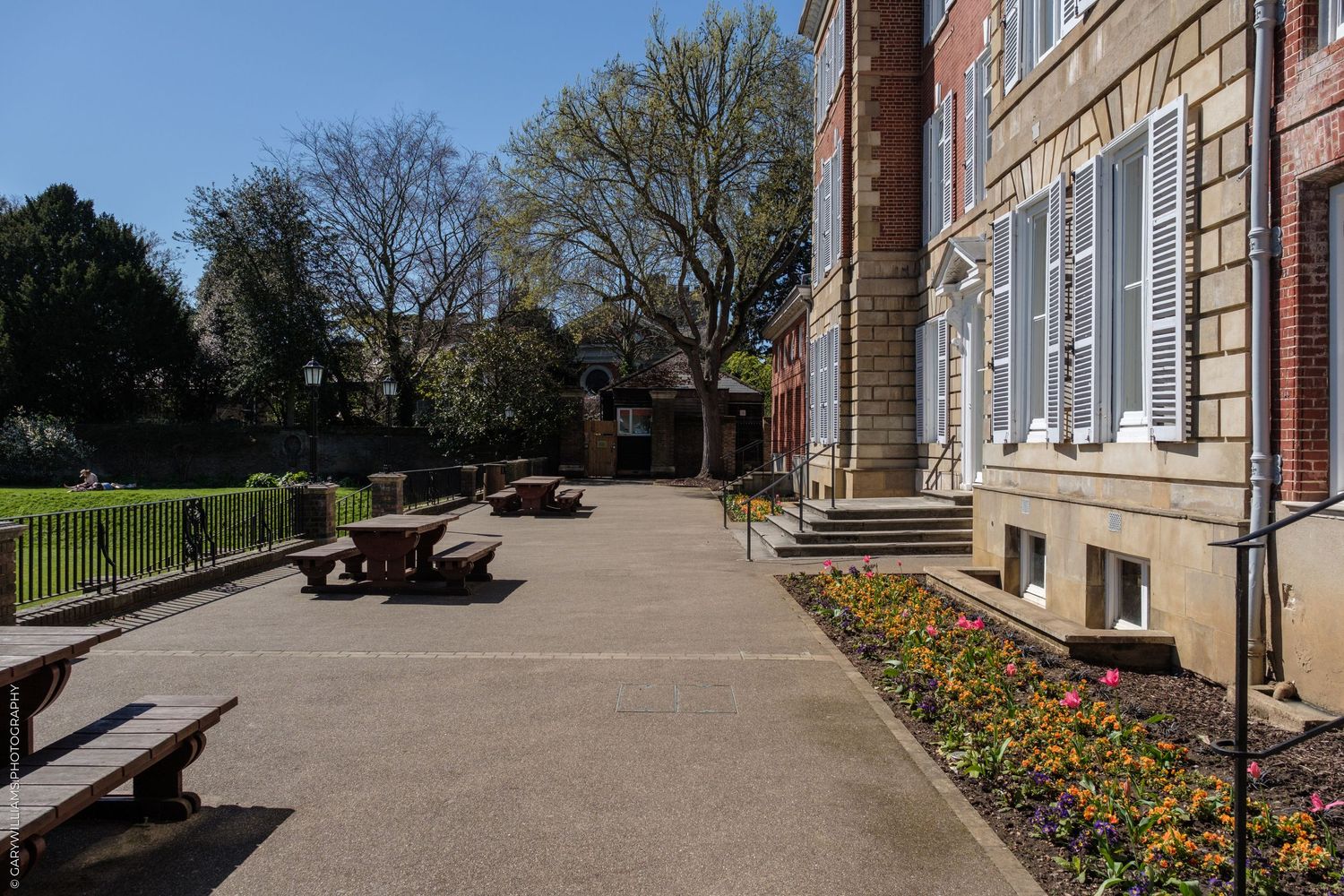 Sunny courtyard of a historic brick building with picnic tables, colorful flower beds, and lush green trees on a clear day.