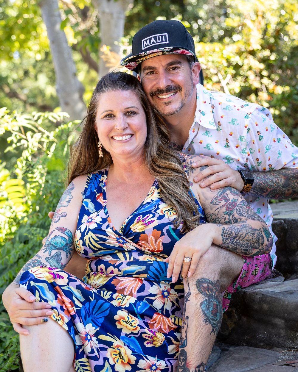 A smiling couple with tattoos sits on stone steps outdoors, wearing tropical outfits, surrounded by lush green trees.
