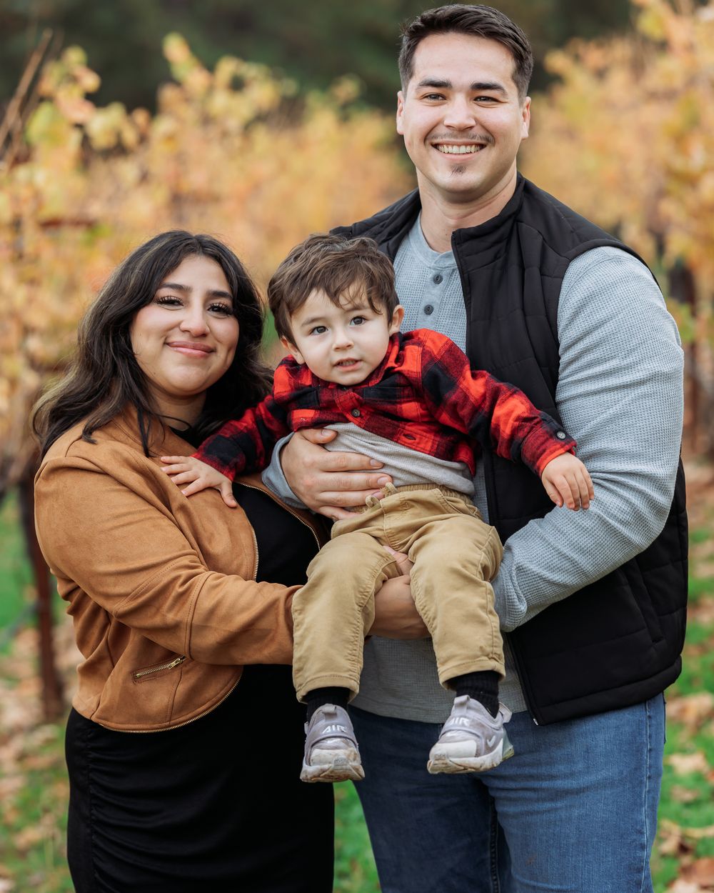 Young couple holding toddler in red plaid jacket, smiling in autumn vineyard setting.
