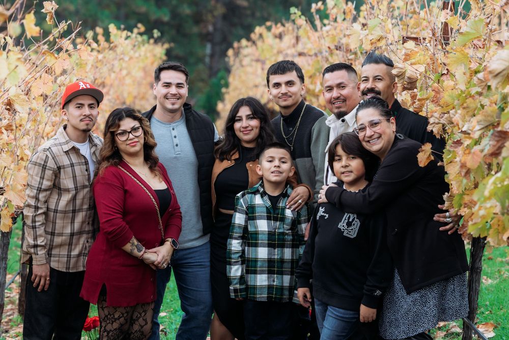 Large Hispanic family posing together in autumn vineyard, smiling outdoors among golden fall foliage.