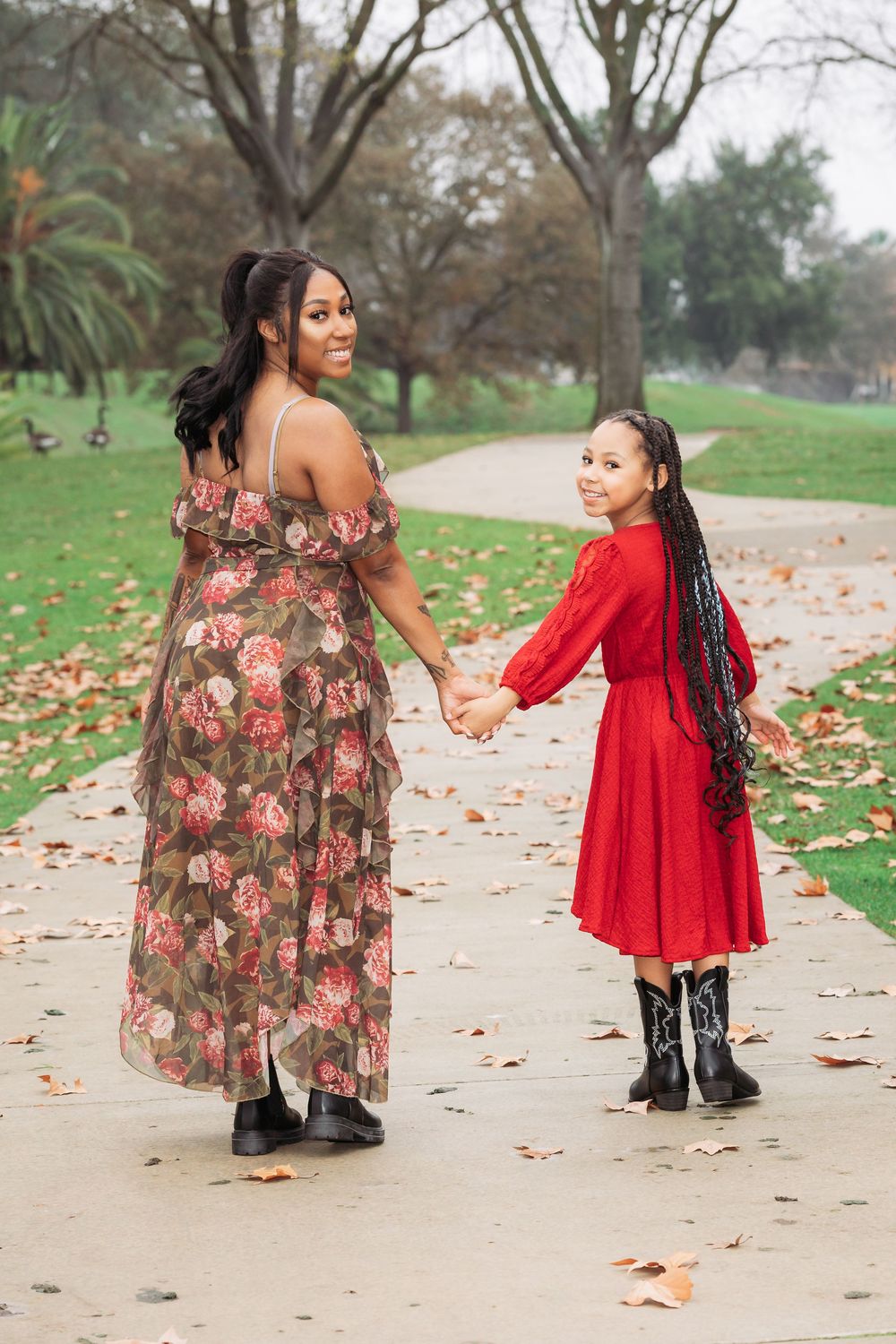 A Black mother and daughter holding hands on a park path, wearing floral and red dresses with boots among autumn leaves.