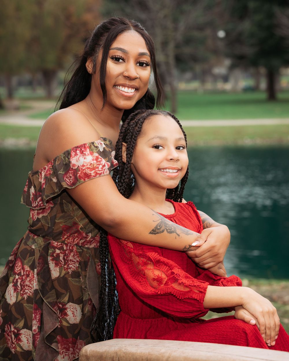 A smiling Black mother hugging her daughter in red dress, posing near a pond in a park for a portrait photo.