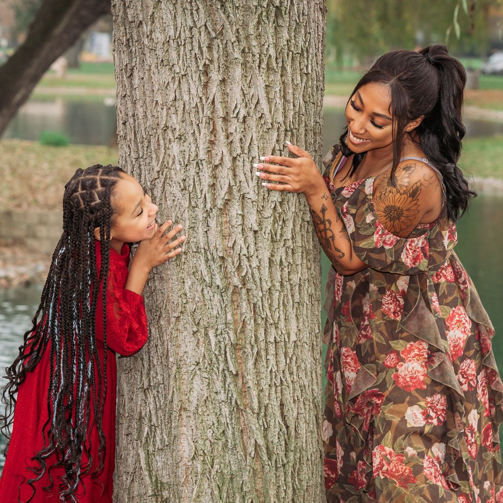 Two girls in colorful dresses peek around a large tree trunk outdoors near a pond.