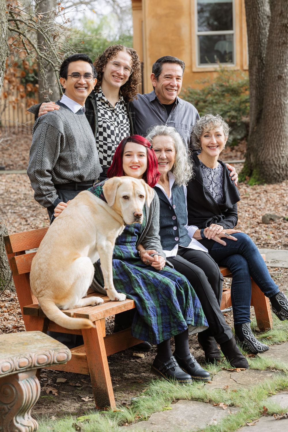 Multi-generational family of six poses outdoors on a wooden bench with a yellow Labrador Retriever in a garden setting.