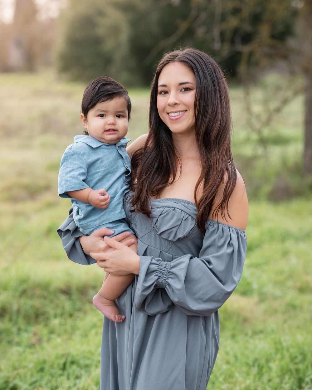 A smiling young woman in a gray off-shoulder dress holds a baby boy in a denim shirt outdoors in a green field.