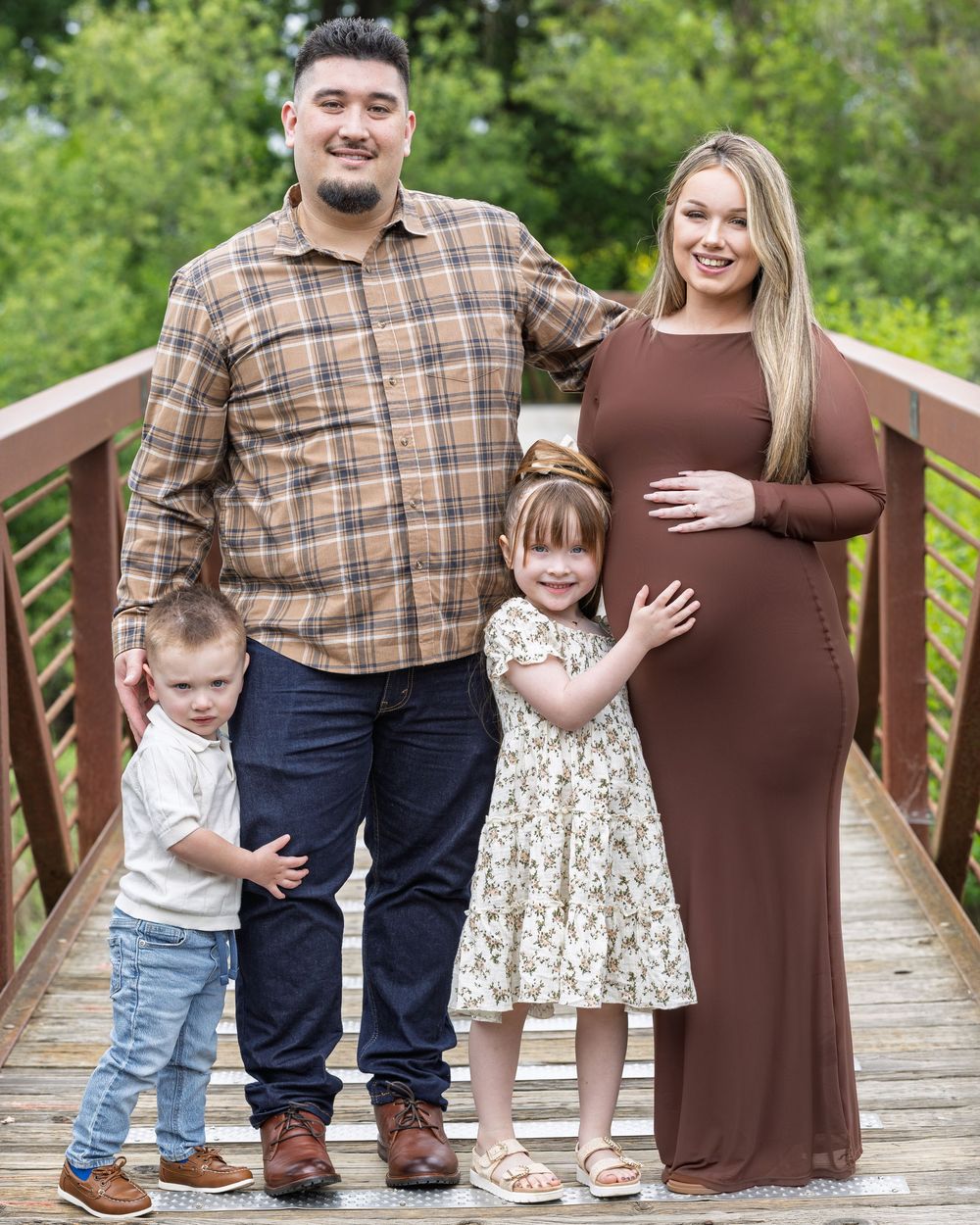 Family maternity photo on wooden bridge, pregnant woman in brown dress with husband and two young children smiling outdoors.