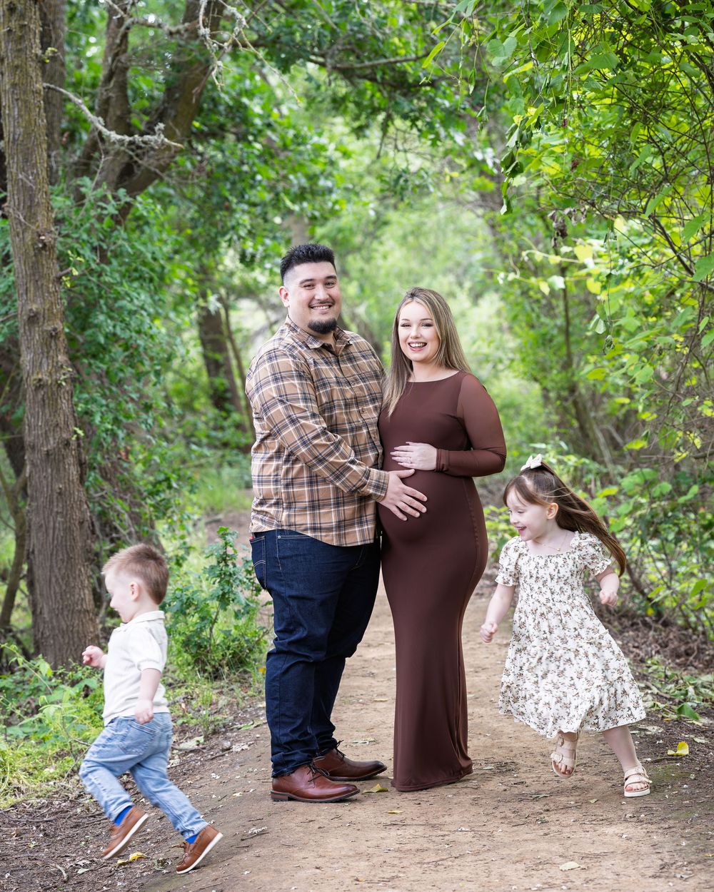 Pregnant woman in brown dress poses with partner and two young children on a scenic woodland path.
