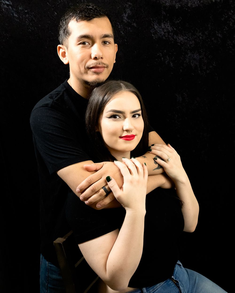 A couple poses intimately against a dark background, the man embracing the woman from behind, both wearing black.