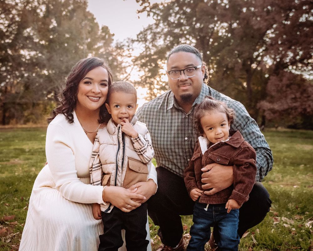 Family of four poses outdoors in autumn park, parents holding two toddlers, warm golden sunlight filtering through trees.