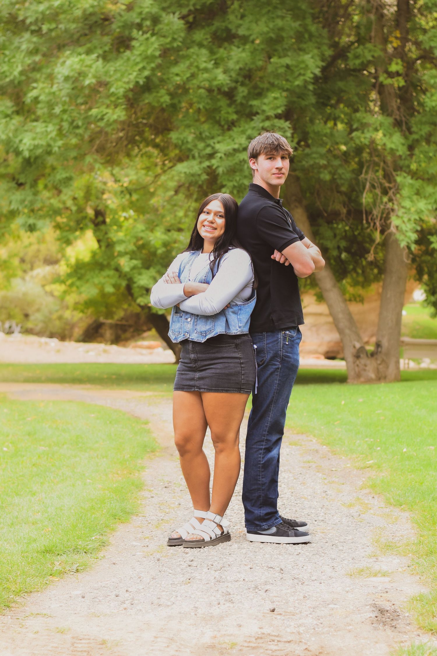 A young couple stands back-to-back with arms crossed on a park path, surrounded by lush green trees.