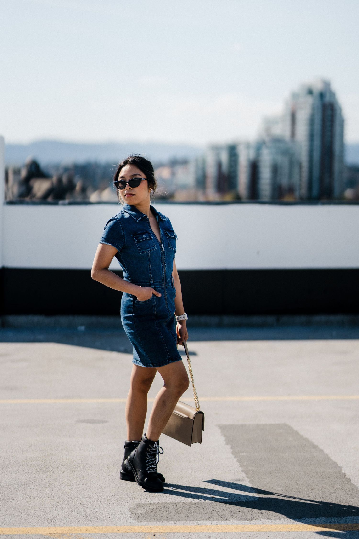 Stylish woman in denim mini dress and black boots holding a small bag on a rooftop with city skyline behind.