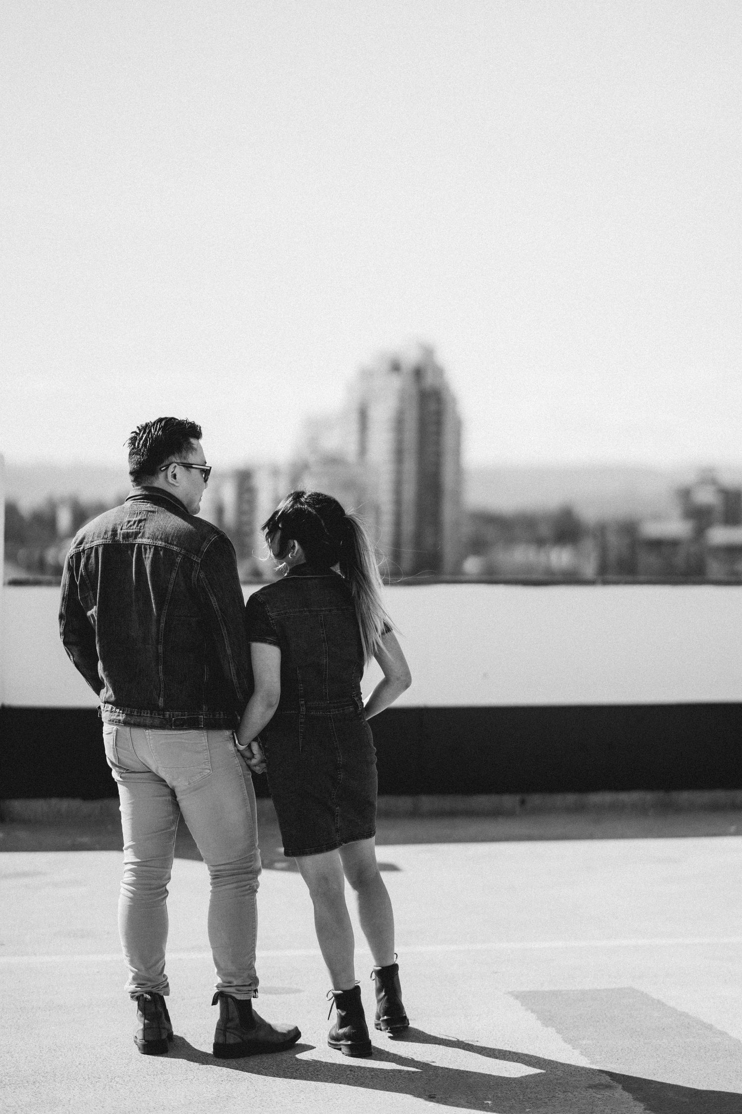 Black and white photo of couple holding hands on rooftop gazing at blurred city skyline in background.