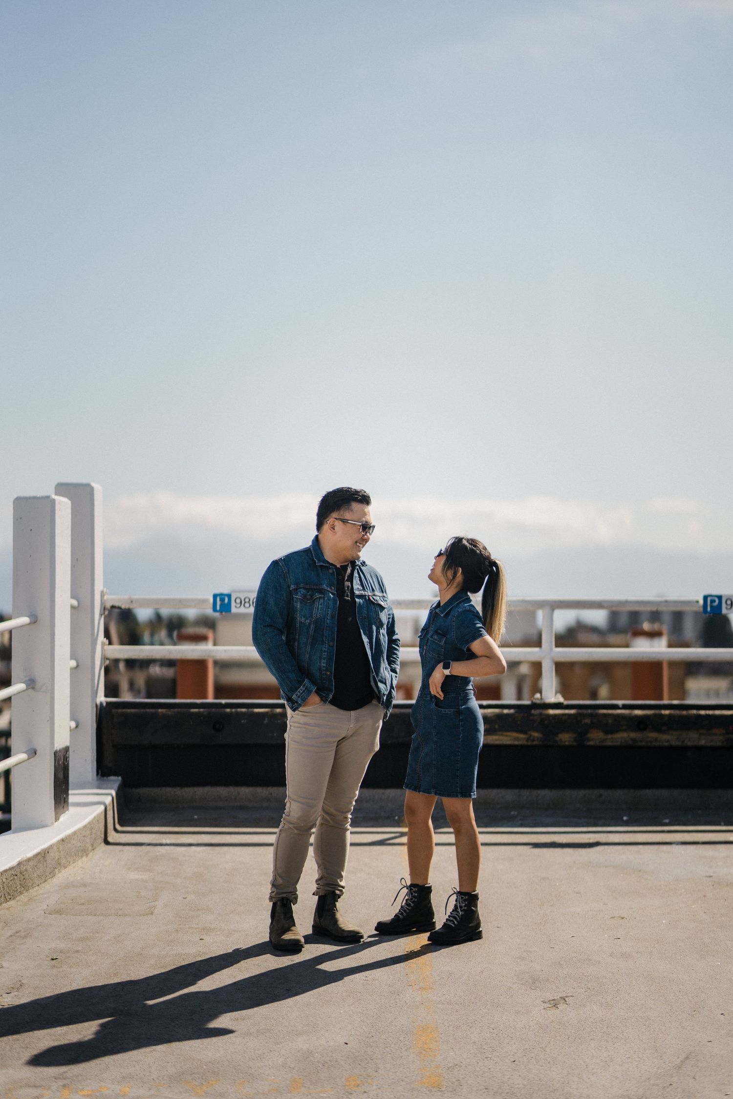 Couple in matching denim outfits and sunglasses facing each other on a rooftop parking lot under a clear blue sky.