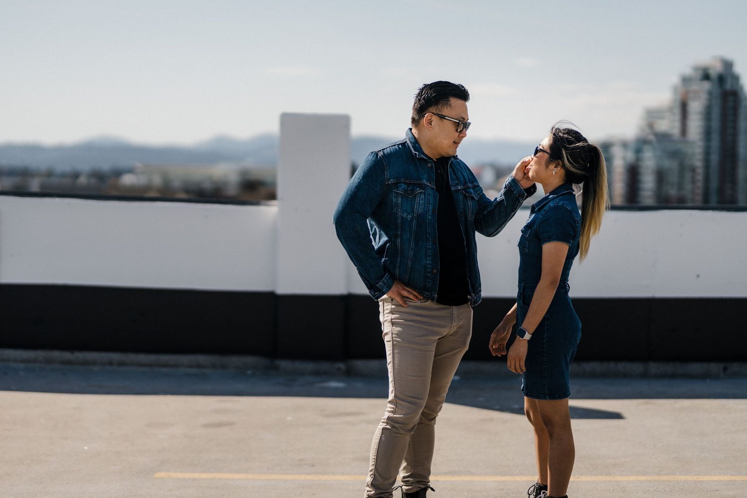 A couple in matching denim outfits stand facing each other on a rooftop with a city skyline backdrop.
