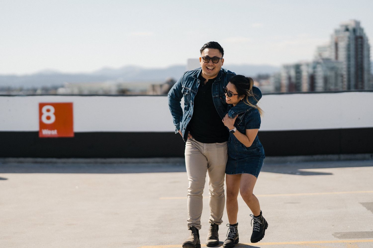 Couple in matching denim outfits posing on rooftop parking with city skyline backdrop.