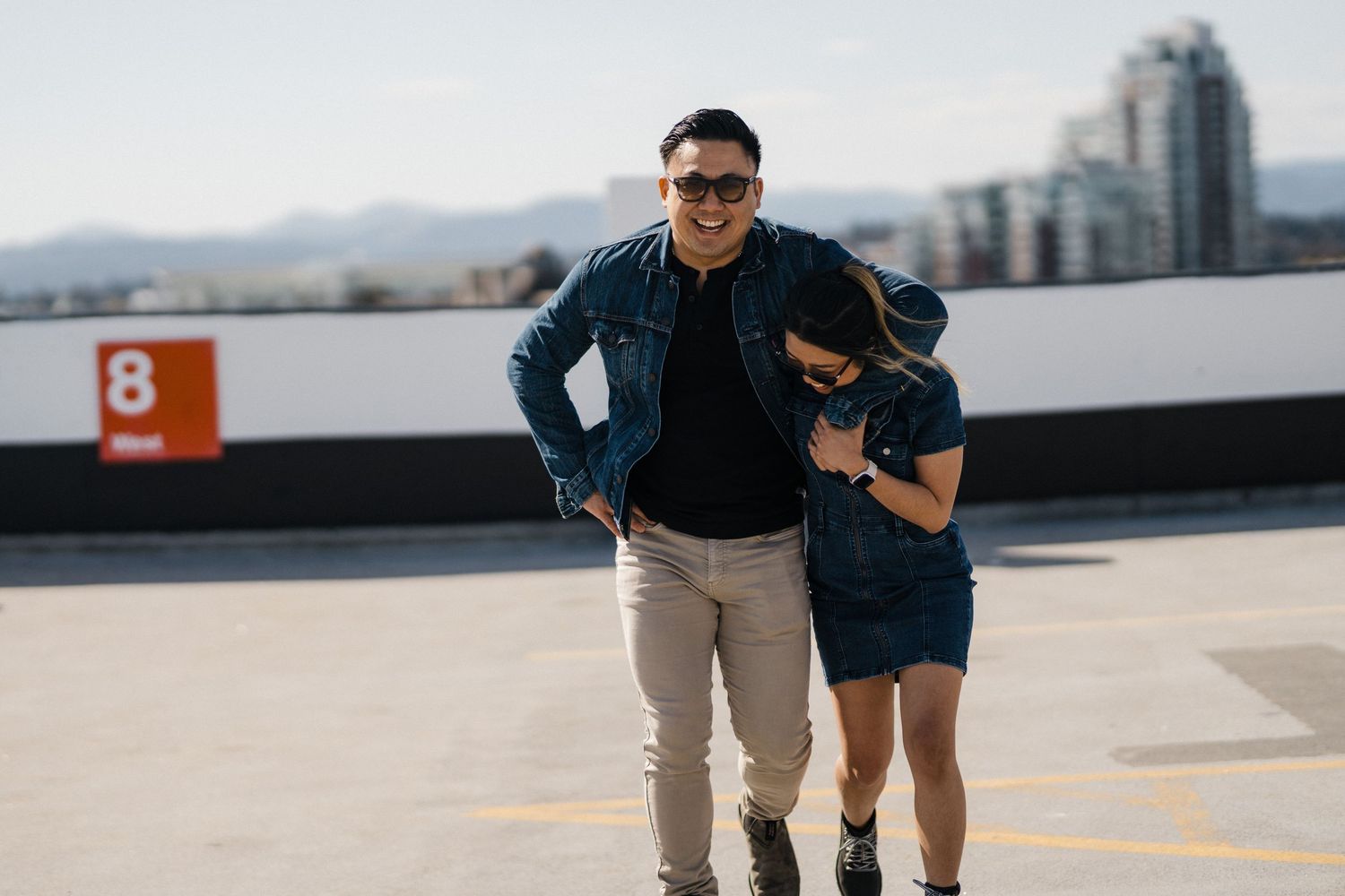 Two people in matching denim outfits pose on a rooftop parking lot with city skyline in the background.
