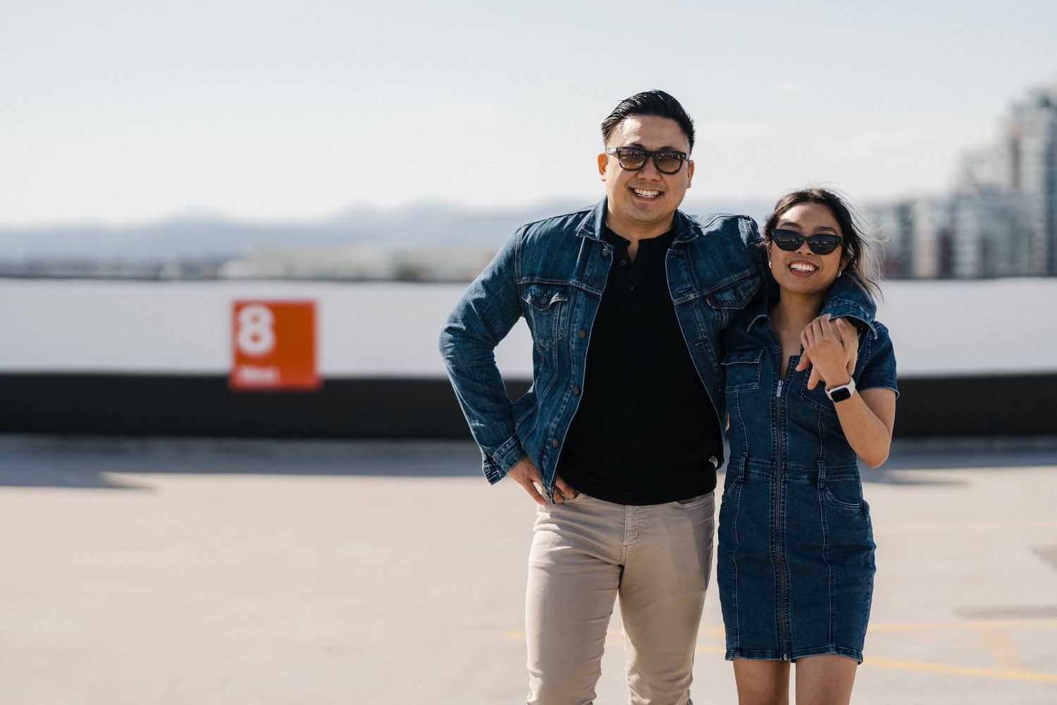 A stylish couple wearing matching denim jackets and sunglasses posing on a rooftop parking lot with a city skyline backdrop.