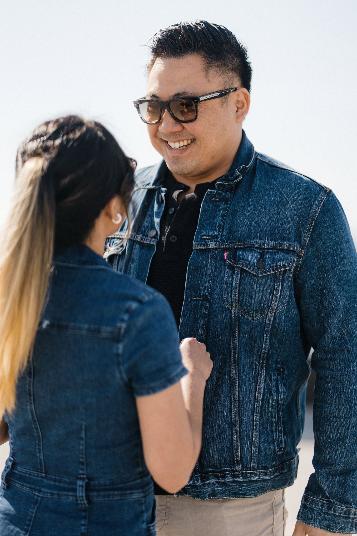 Smiling man in denim jacket and sunglasses facing woman in denim dress outdoors.