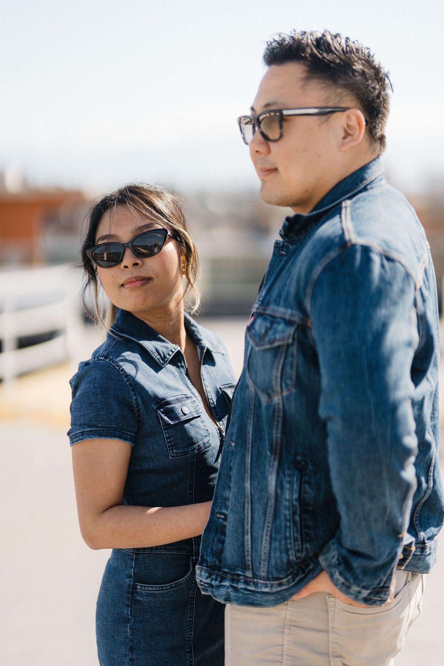 Asian couple in coordinating denim jackets posing outdoors, woman in sunglasses looking at camera.