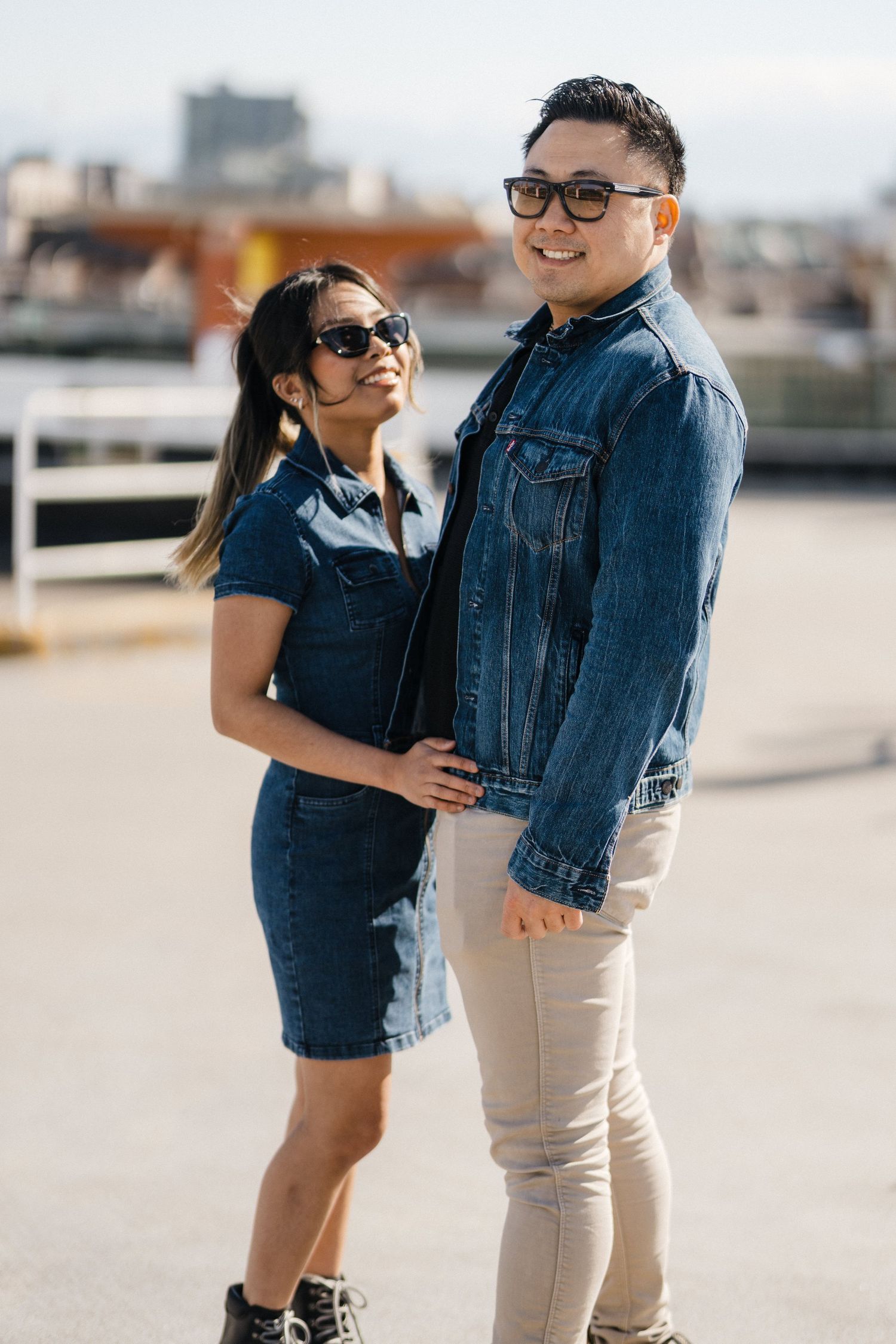 Smiling Asian couple in coordinated denim outfits and sunglasses posing together on a sunny rooftop parking area.