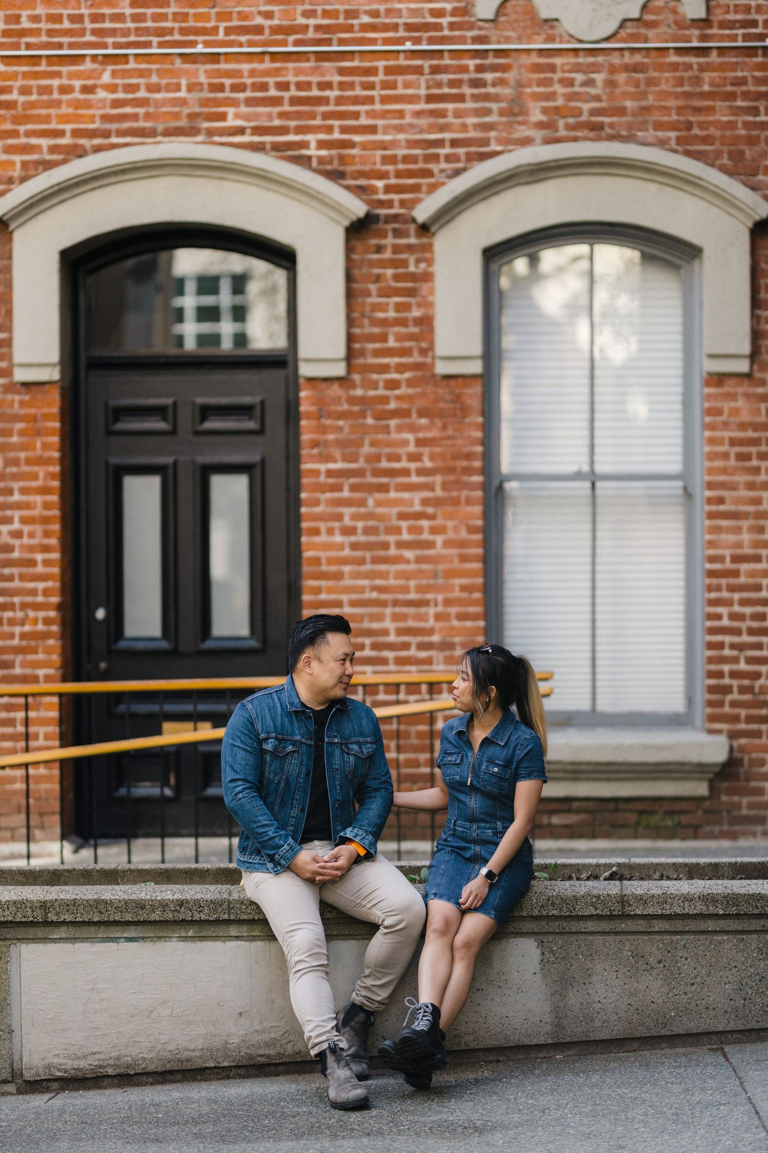 A couple in matching denim jackets sit on steps outside a red brick building, sharing a warm conversation.