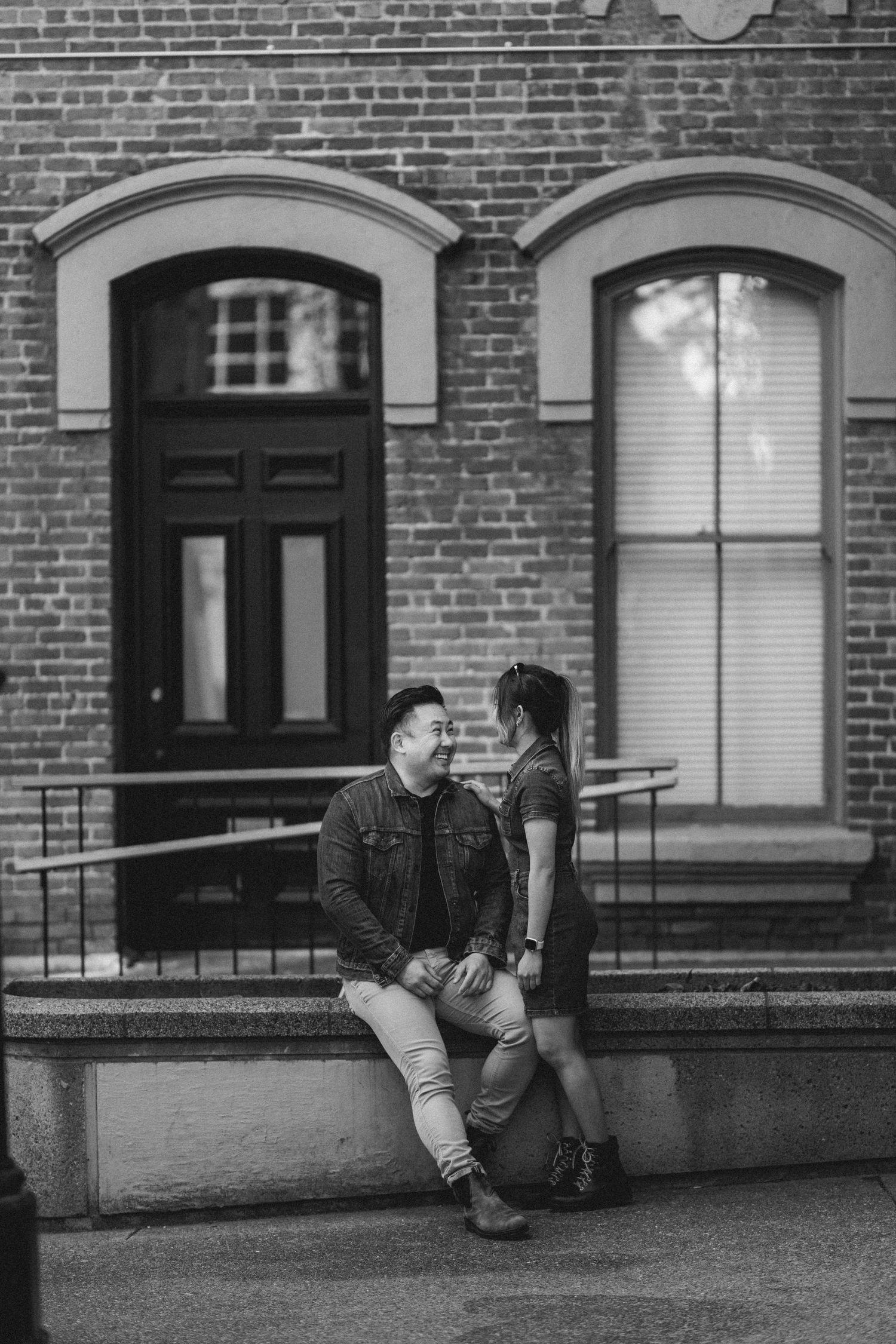 Black and white photo of a couple sitting and standing on steps outside a brick building, sharing a tender moment.