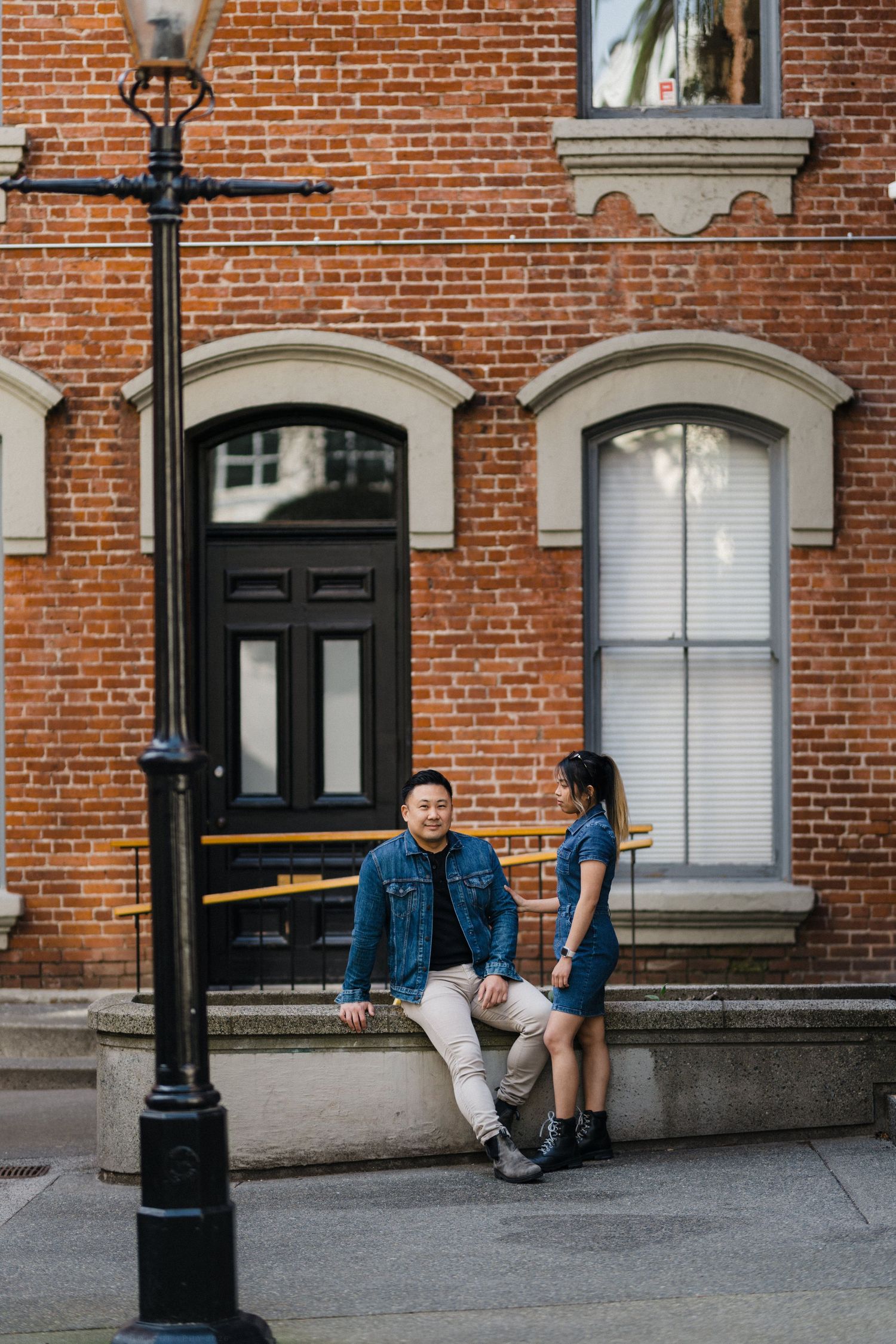 A couple relaxes outside a historic red brick building, the man seated as the woman stands beside him smiling.