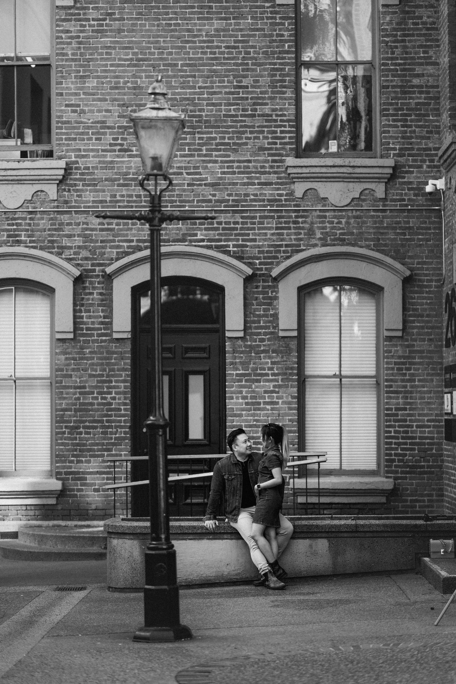 Black and white photo of a couple sitting together by a lamppost outside a brick building.