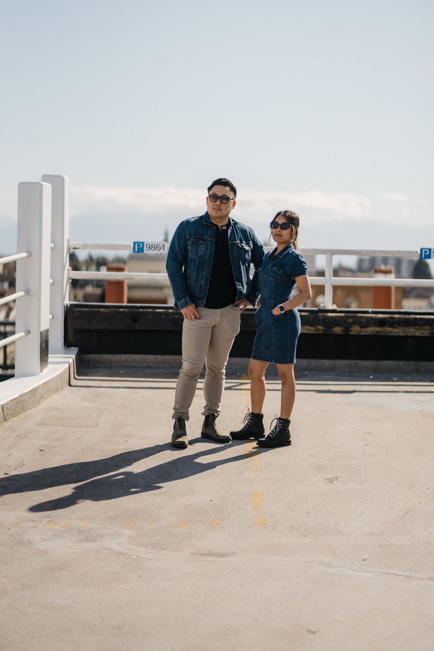 Couple in matching denim outfits and sunglasses posing on a rooftop parking lot on a sunny day.