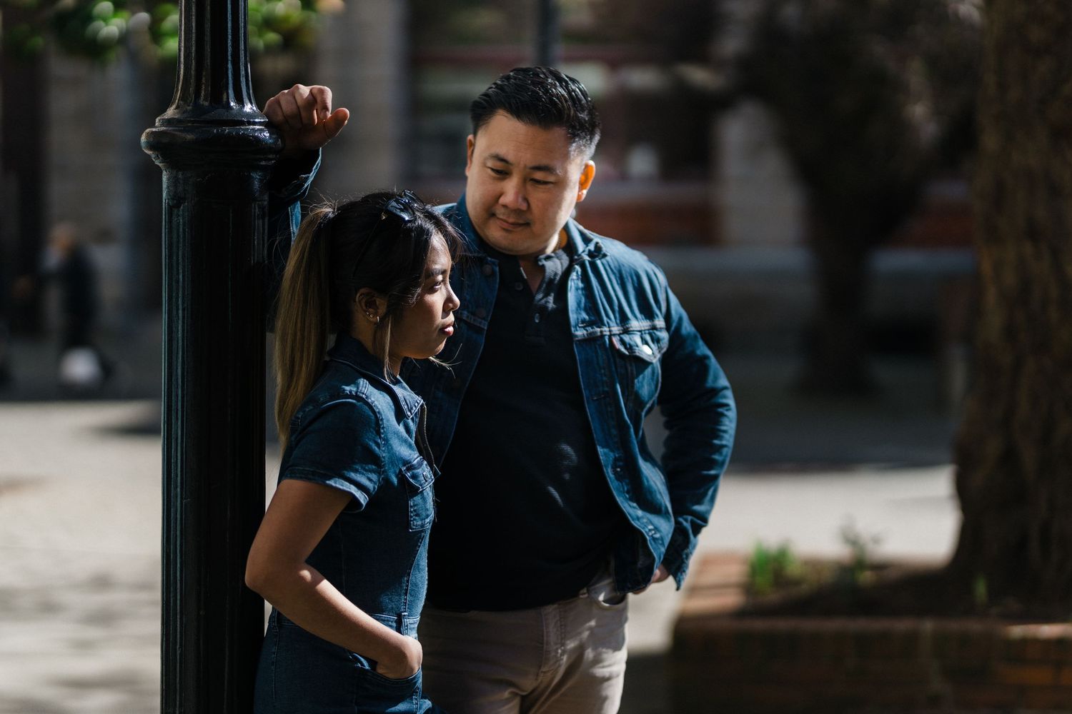 Asian couple leaning against a black lamppost in an urban park, wearing coordinated denim outfits.