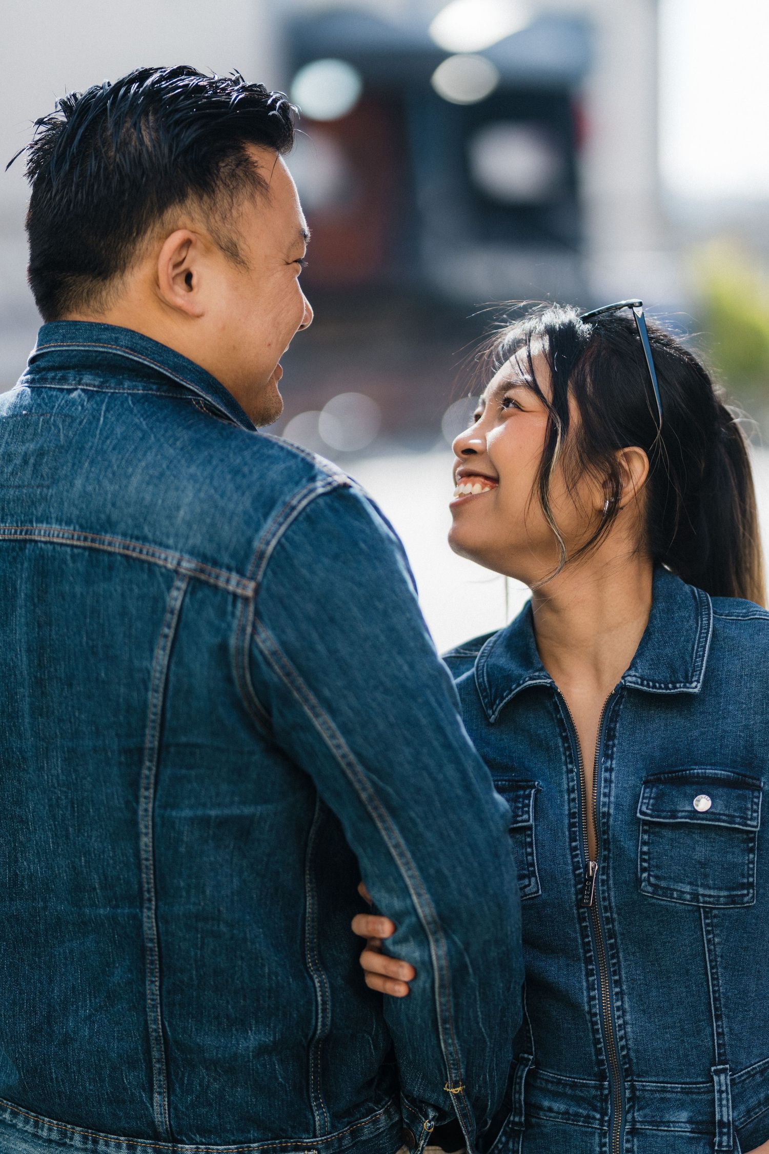 A smiling Asian couple in denim jackets share a tender moment, with the woman gazing up at her partner.