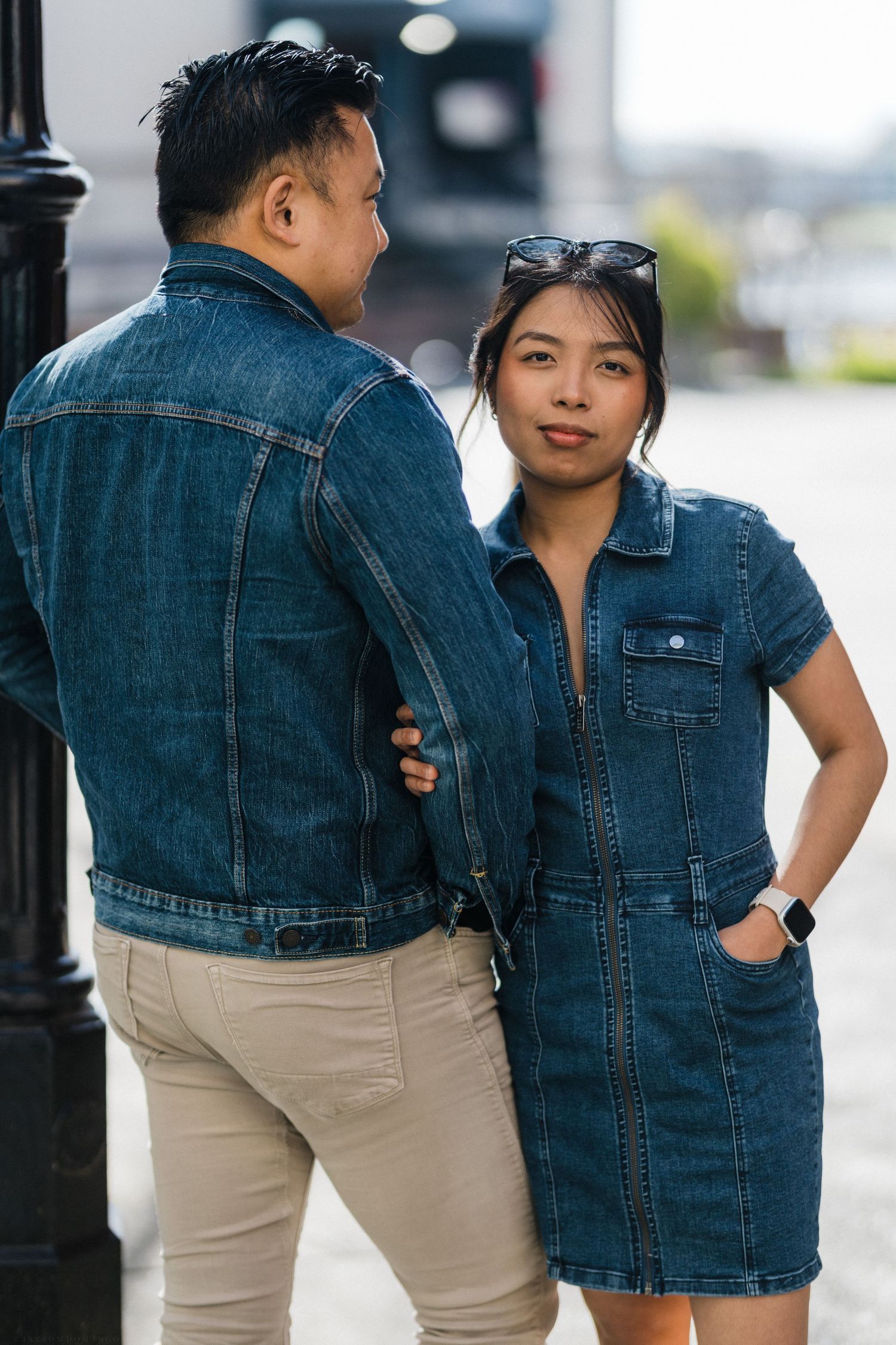 Asian couple in matching denim outfits pose on urban street, woman in denim dress smiles at camera.