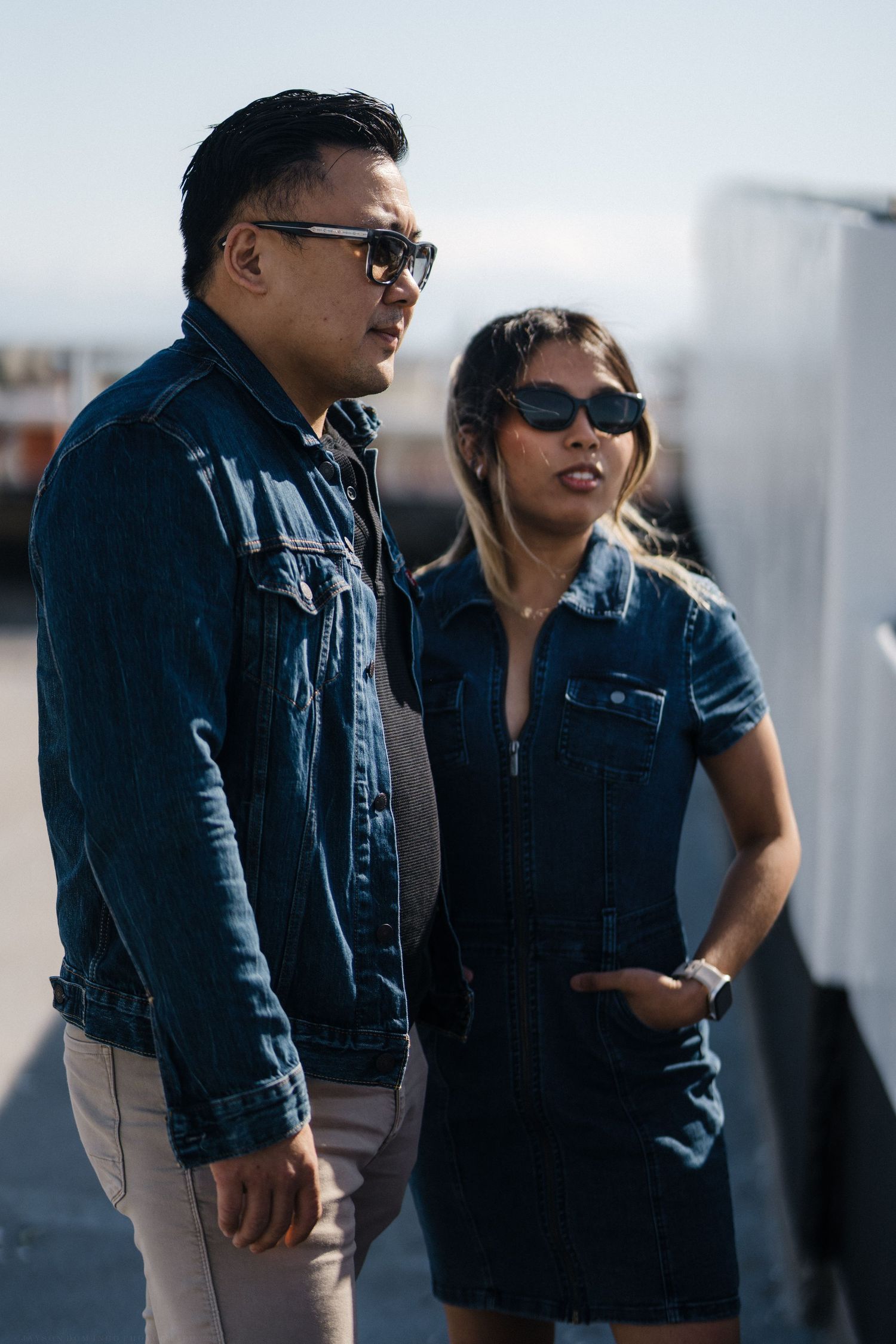 Trendy couple wearing matching denim outfits and sunglasses posing outdoors in sunlight.