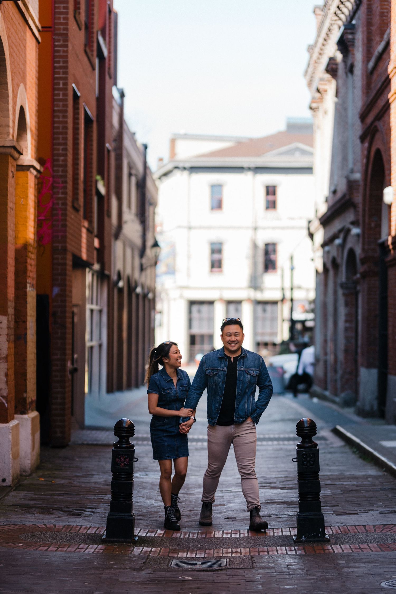 A stylish couple in coordinating denim outfits hold hands while walking through a charming urban brick alleyway.