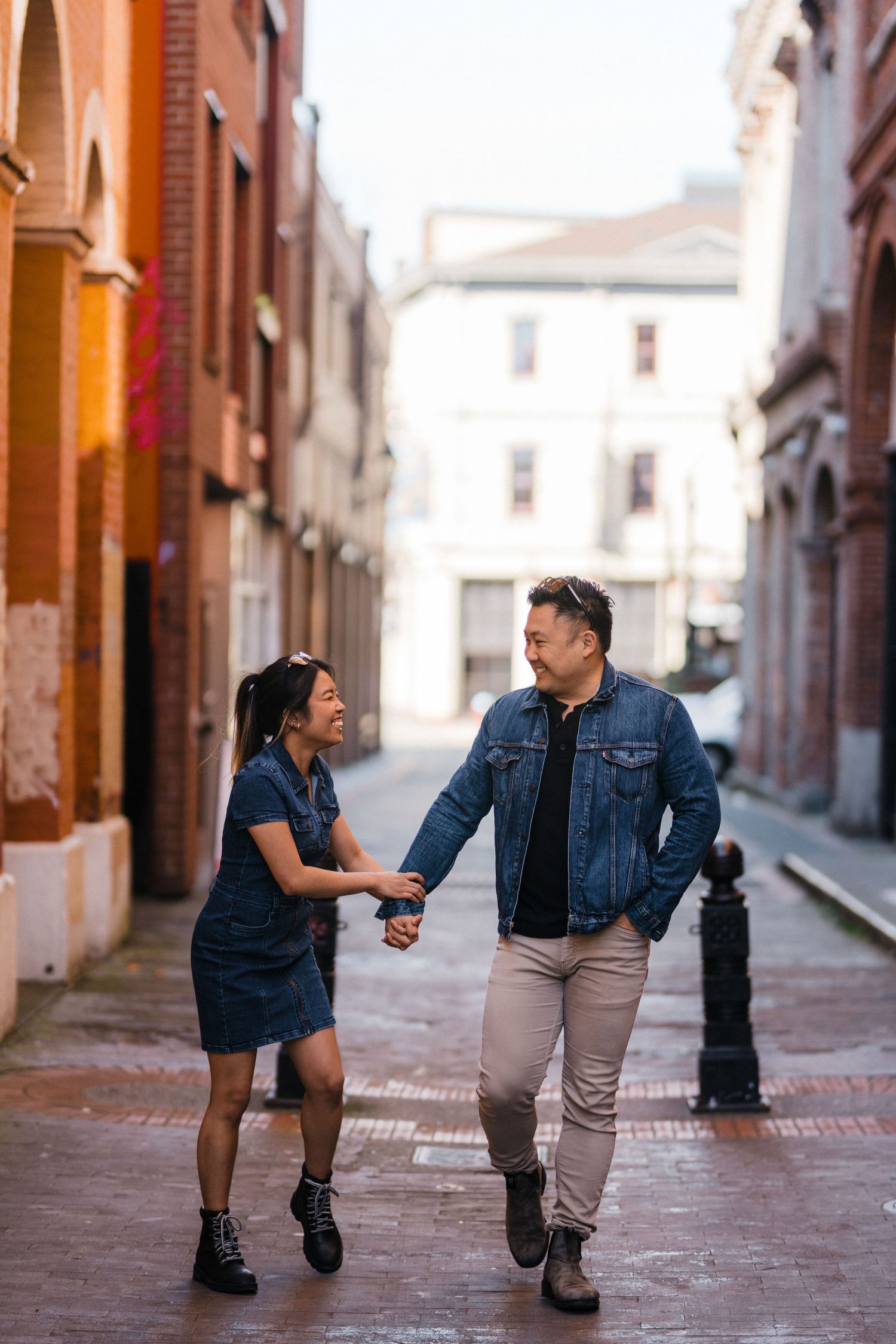 A happy couple in matching denim outfits walk hand-in-hand through a sunlit cobblestone alley between brick buildings.