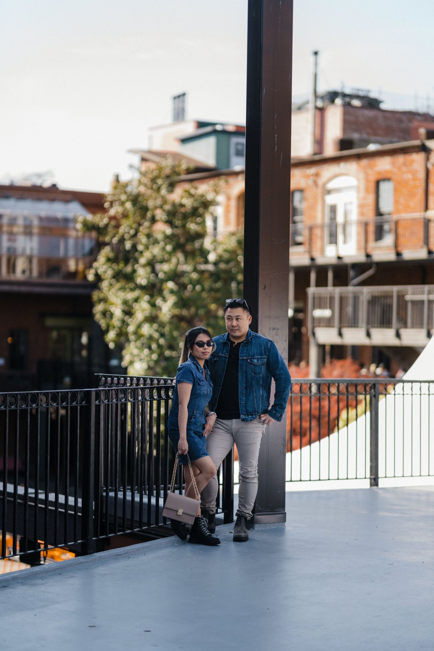 Stylish couple in denim outfits standing on urban bridge with brick buildings behind.