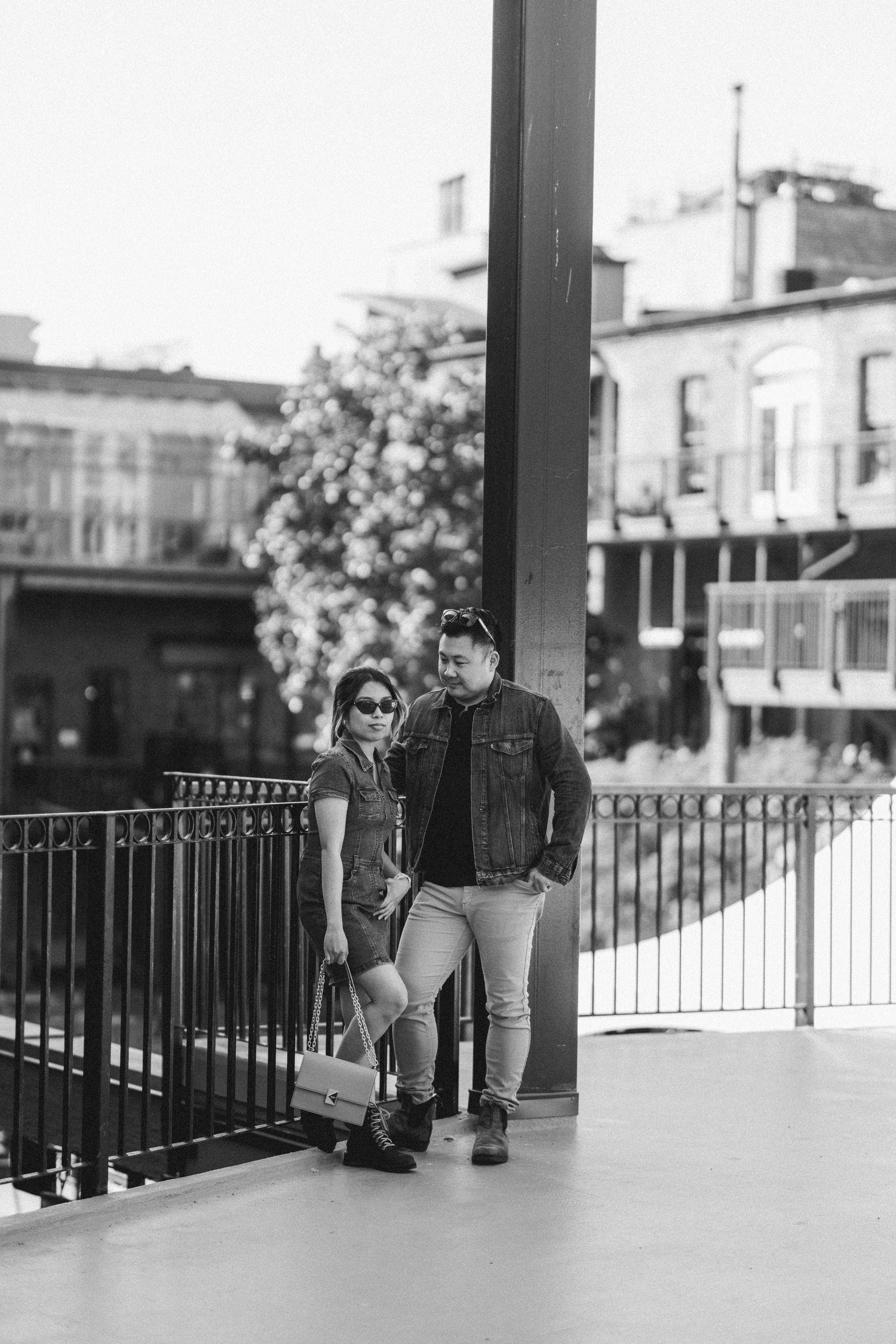 A black and white photo of a couple in denim standing together near a railing in an urban outdoor setting.