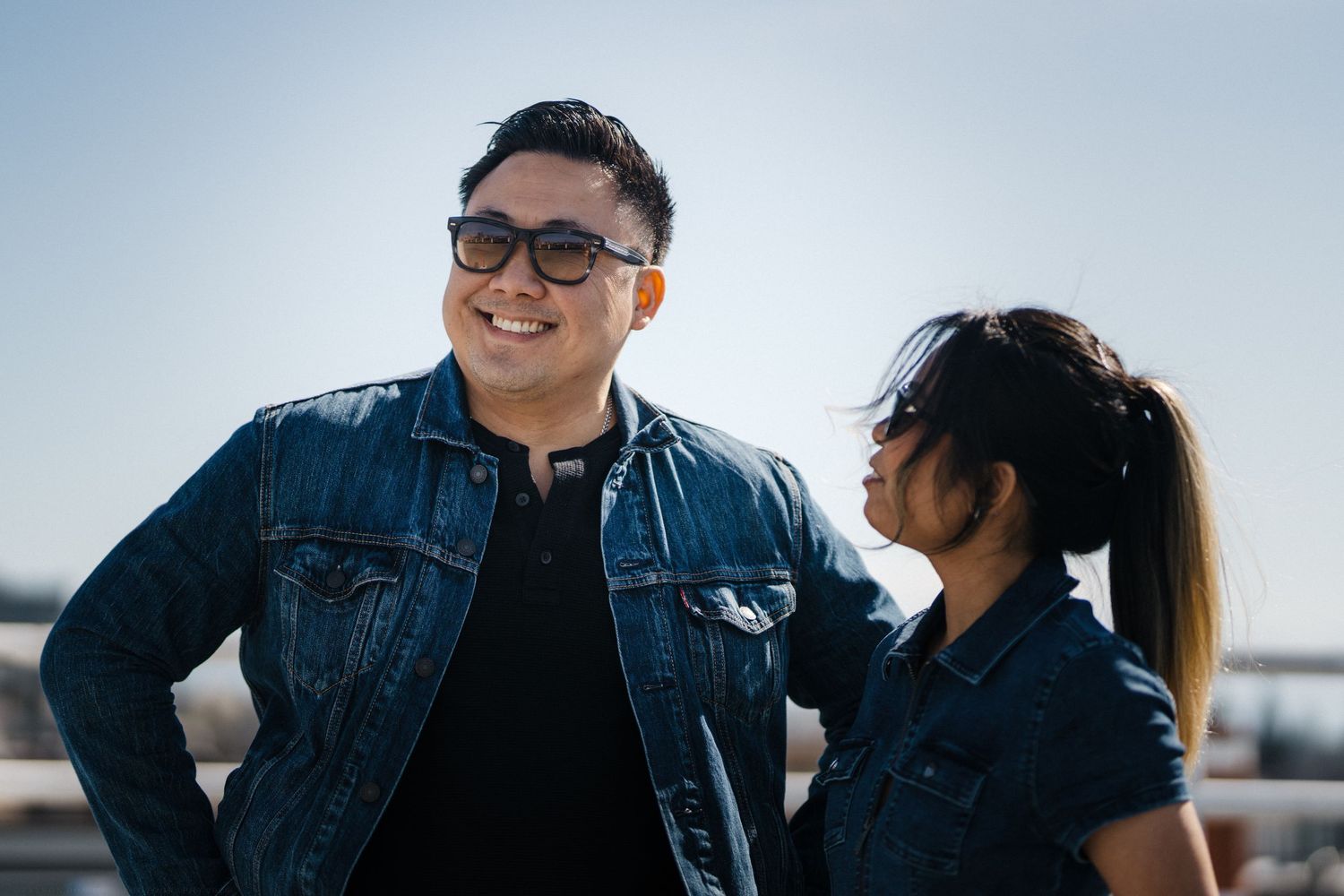Two people wearing matching denim jackets smile outdoors on a sunny day against a bright blue sky.