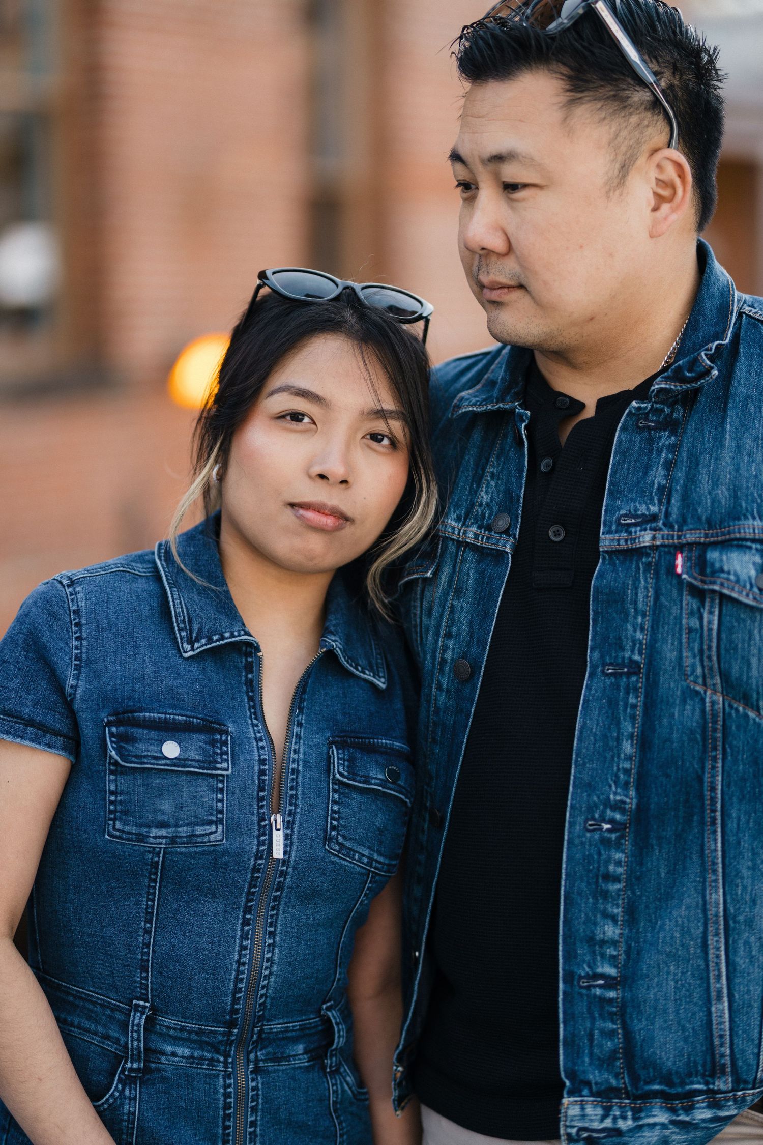 Asian couple in matching denim jackets pose closely together against brick wall backdrop.