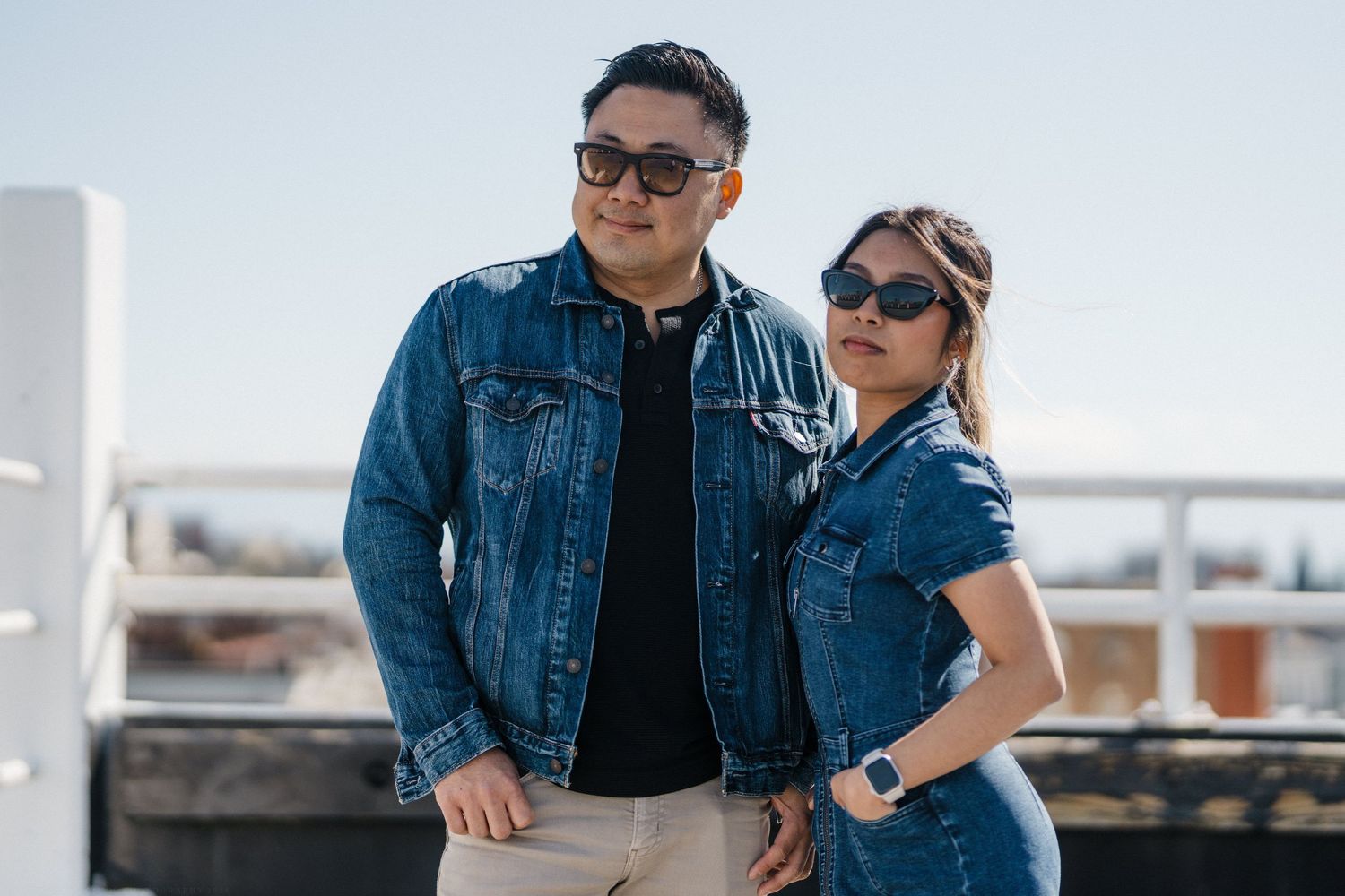 Asian couple wearing coordinating denim jackets and sunglasses posing together on a sunny rooftop location.