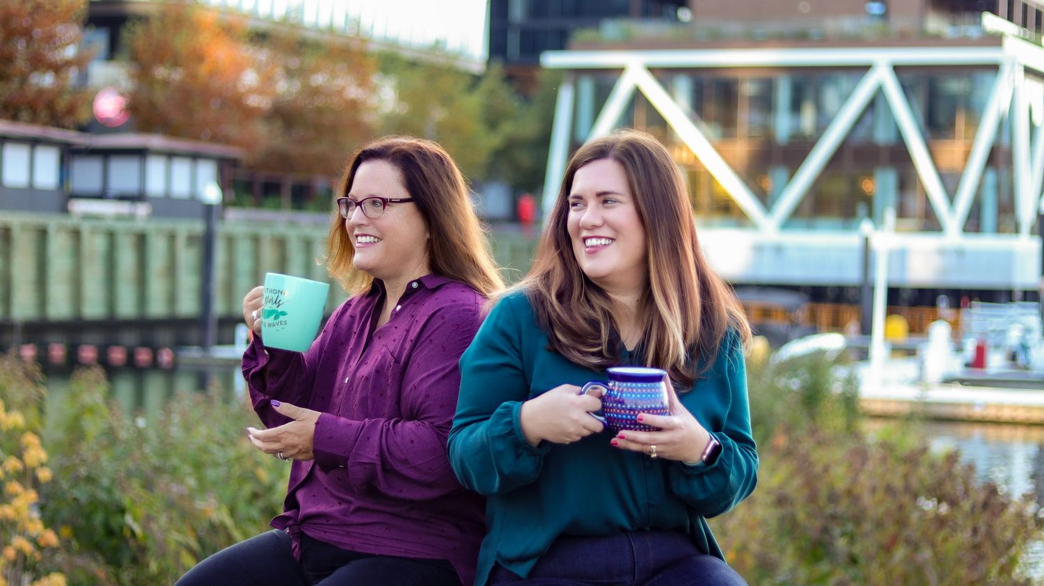 Two women smiling outdoors holding colorful mugs near a waterfront on a autumn day.