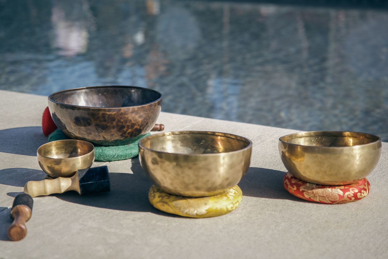 Three golden Tibetan singing bowls on colorful cushions displayed outdoors near a pool with wooden mallets.