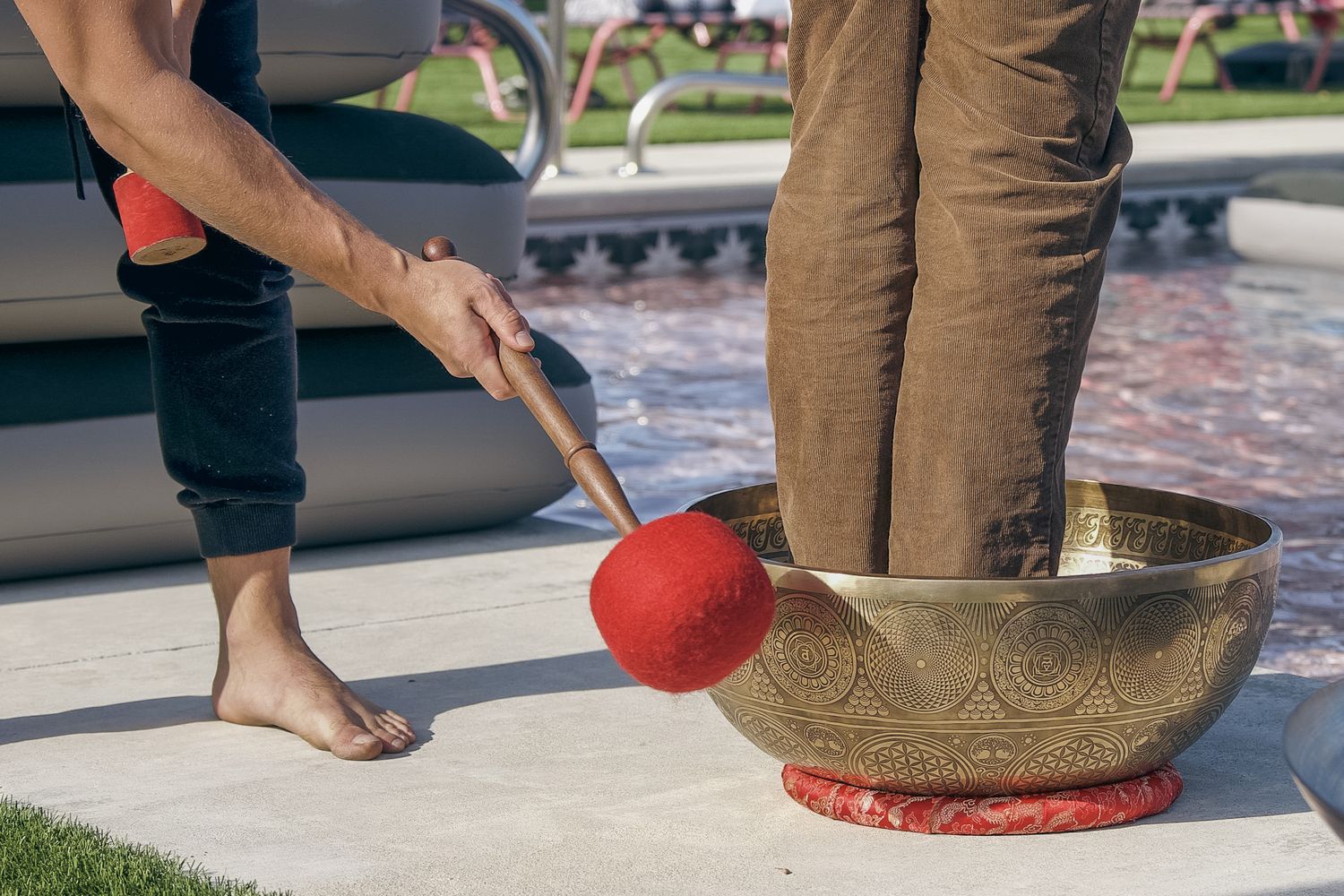 Person playing a Tibetan singing bowl with a red mallet near a pool during an outdoor meditation session.