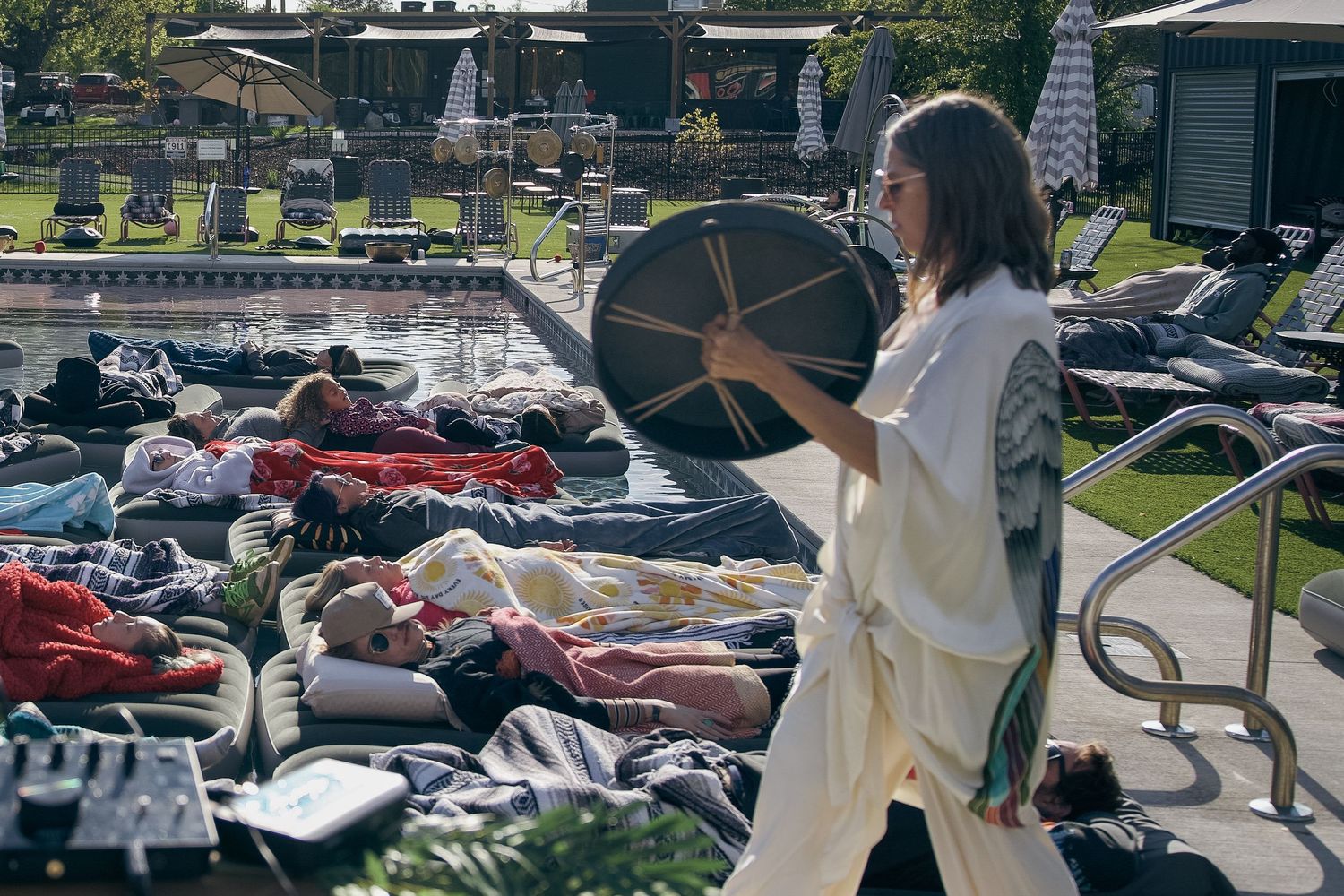 A woman in white linen plays a drum beside a poolside where guests relax on lounge chairs in a sunny resort setting.