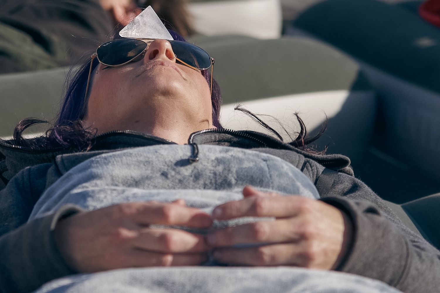 Person lying down relaxing outdoors wearing sunglasses with a paper on forehead, hands folded on chest in sunlight.