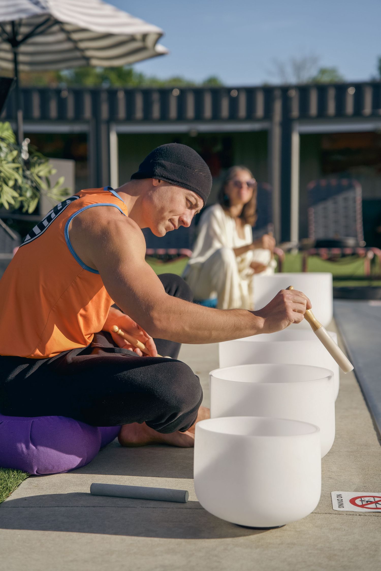 Man in orange tank top playing crystal singing bowls outdoors during a sound healing session.